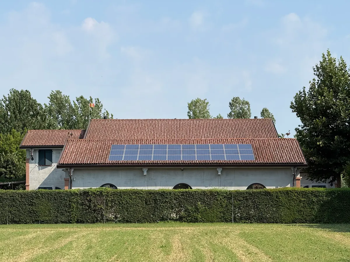 Capannone con tetto in coppi e pannelli fotovoltaici, dietro una siepe e un prato