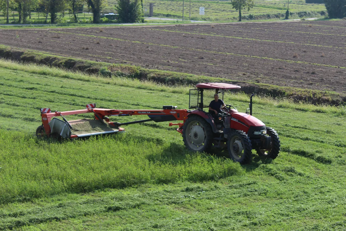 Trattore rosso con la falciatrice a dischi che taglia l’erba medica, con la terra arata e la strada sullo sfondo