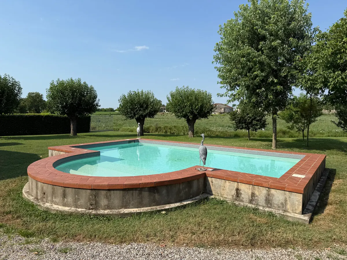 Vasca bordata in cotto con una scultura di airone, tra gli alberi del giardino e i campi