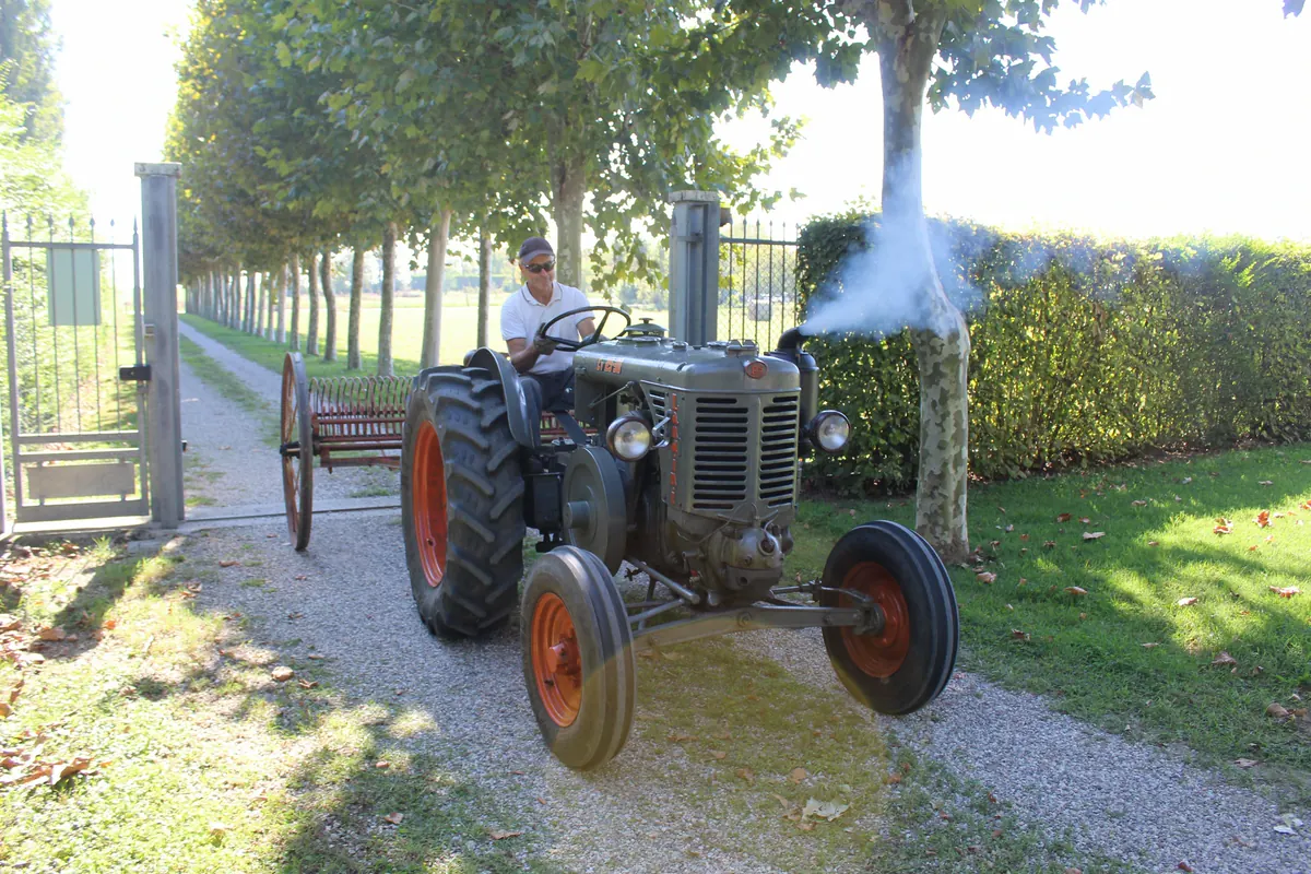 Il trattore esce dal cancello del viale con la rastrellona dietro e uno sbuffo di fumo bianco