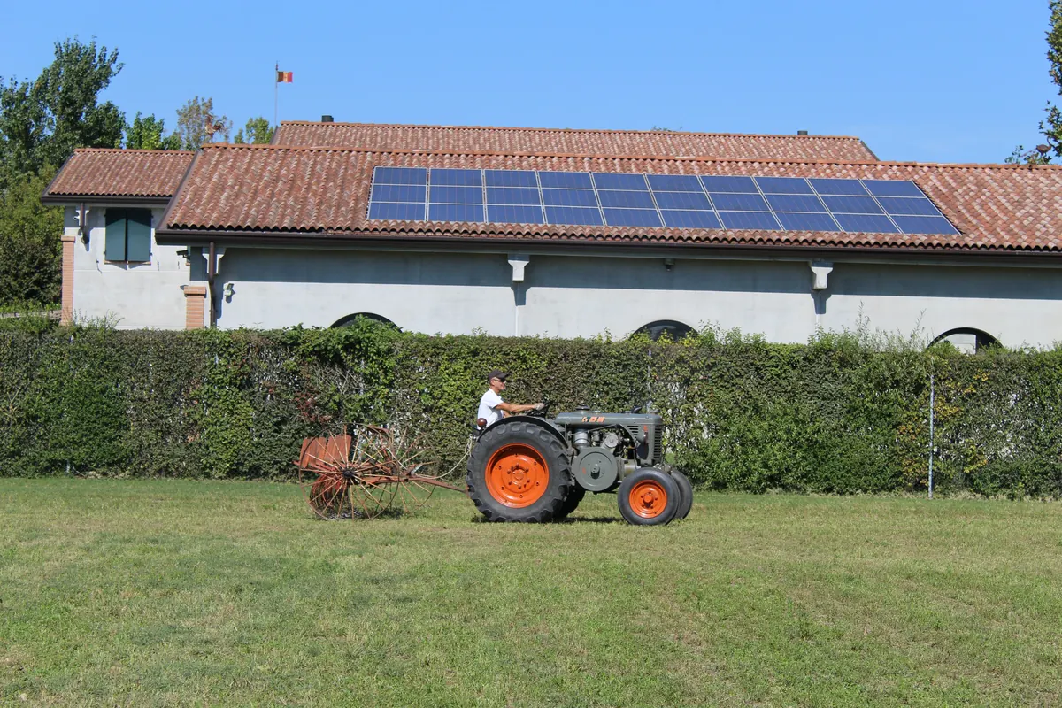 Il trattore con la rastrellona passa lungo la siepe davanti al capannone con i pannelli fotovoltaici sul tetto