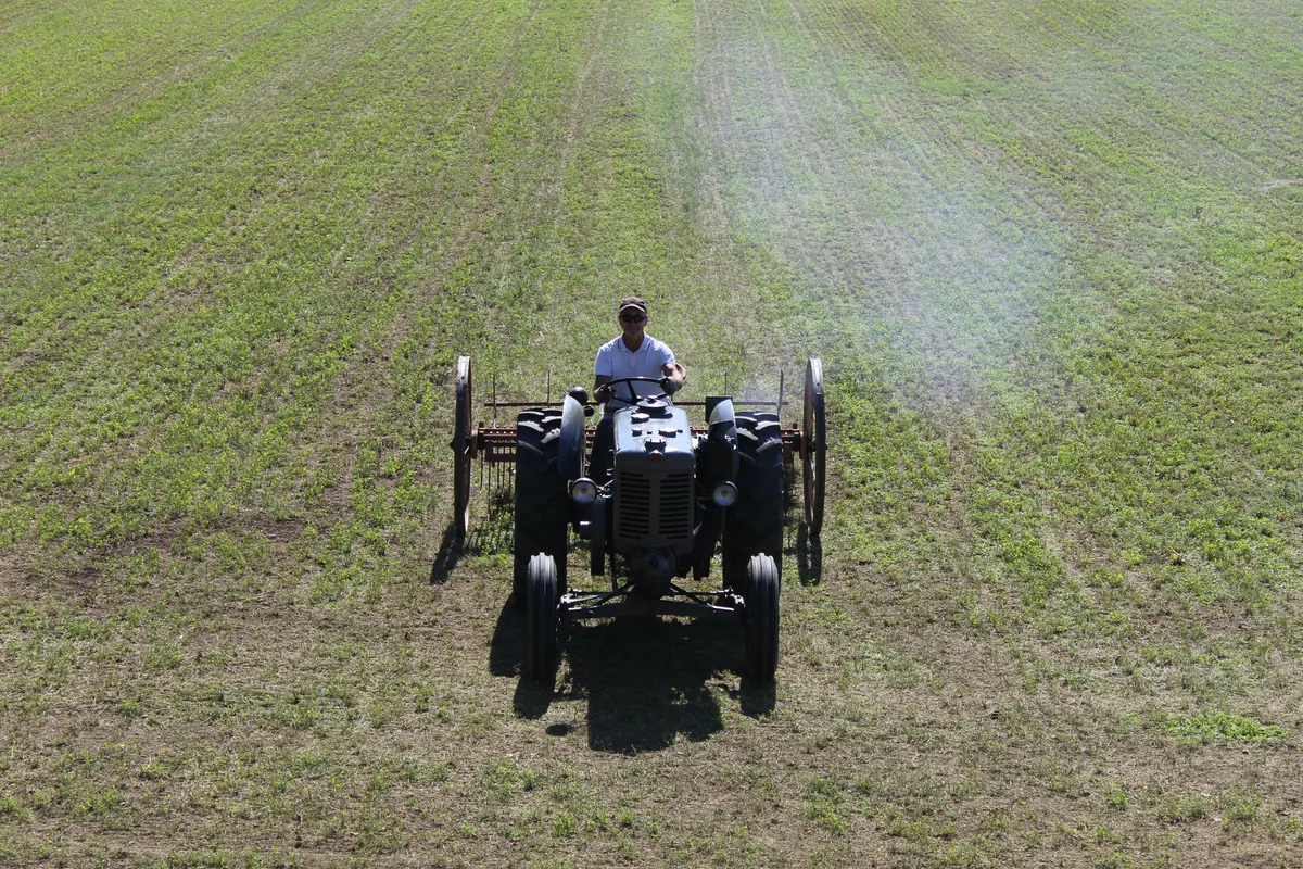 Il vecchio trattore visto di fronte dall’alto in mezzo al campo, con la rastrellona dietro e uno sbuffo di fumo bianco