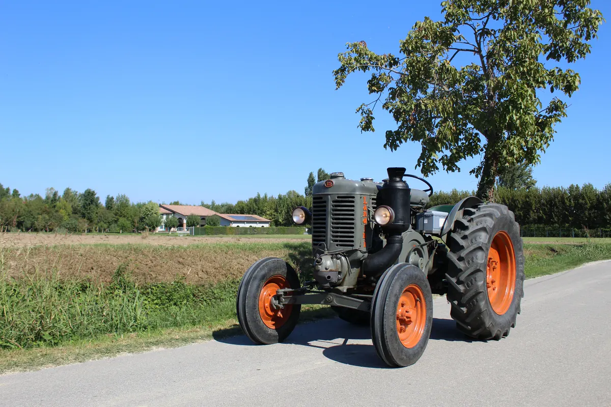 Il vecchio trattore sulla strada asfaltata tra i campi, con un albero e il cielo azzurro