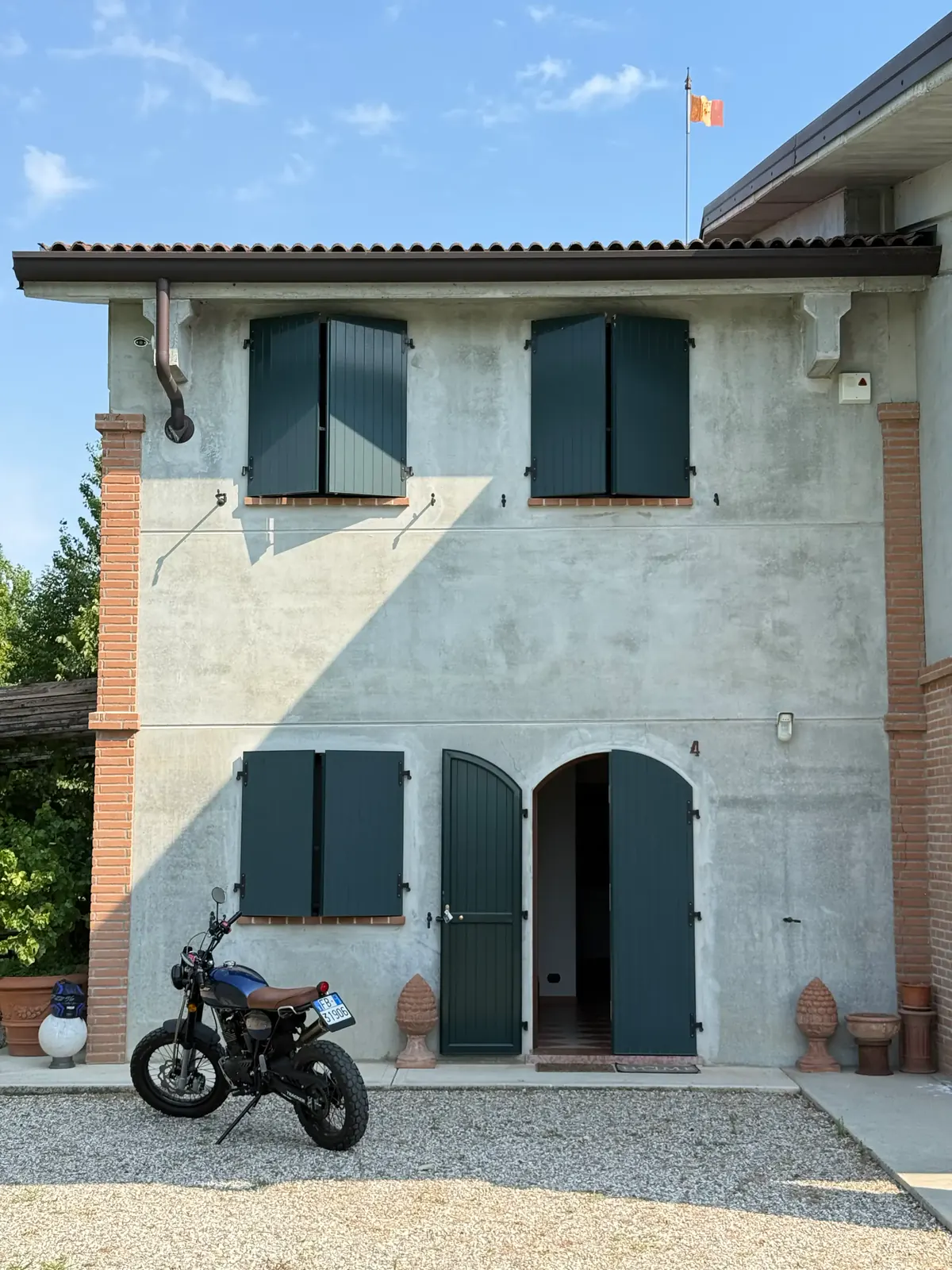 Facciata della cascina con persiane verdi, pilastri in mattone e una moto parcheggiata sul ghiaino