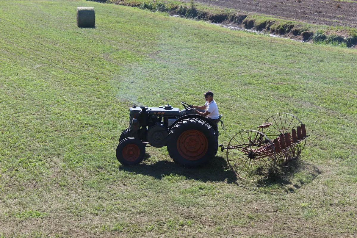 Il trattore con la rastrellona nel campo, con una rotoballa rimasta sull’erba e la terra arata sullo sfondo