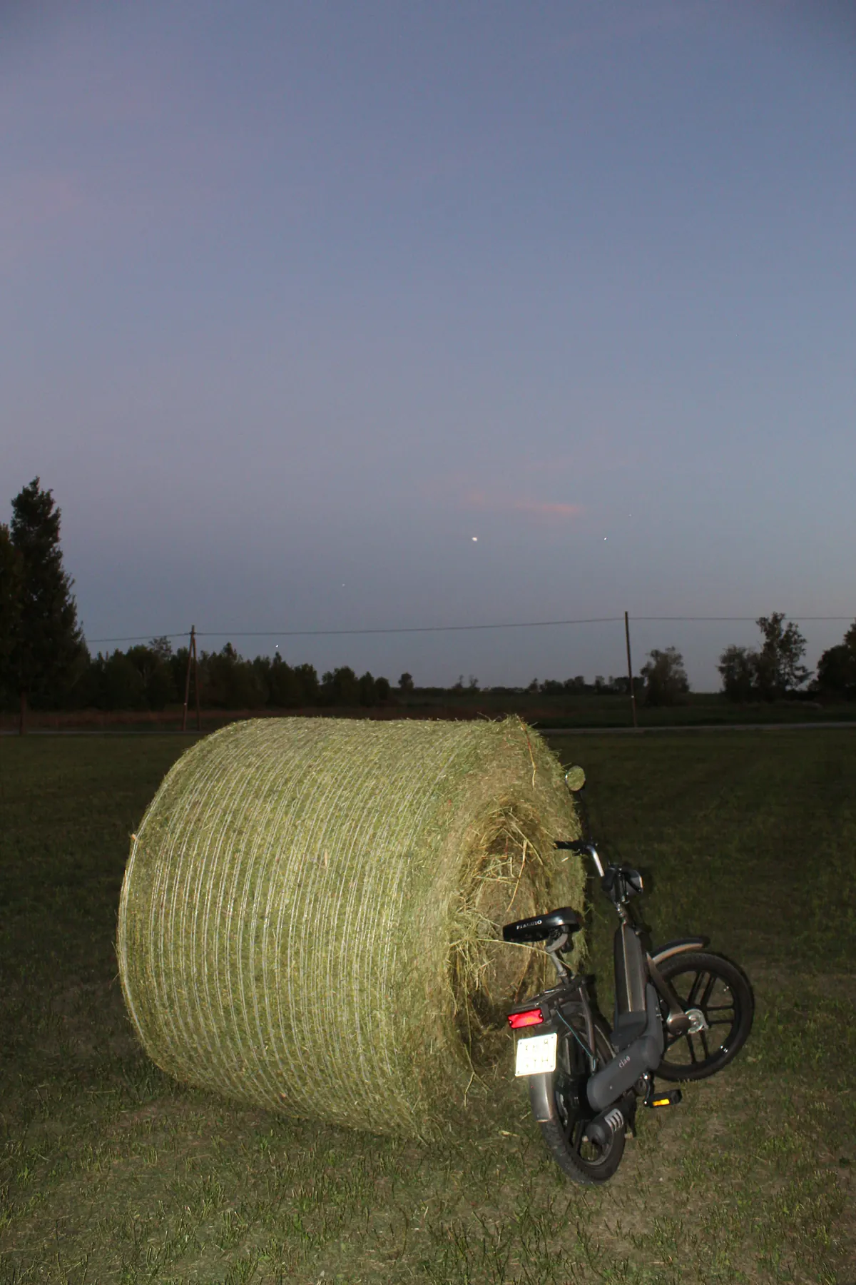 Una rotoballa di fieno nel campo al crepuscolo con il motorino appoggiato dietro, sotto un cielo viola con una stella