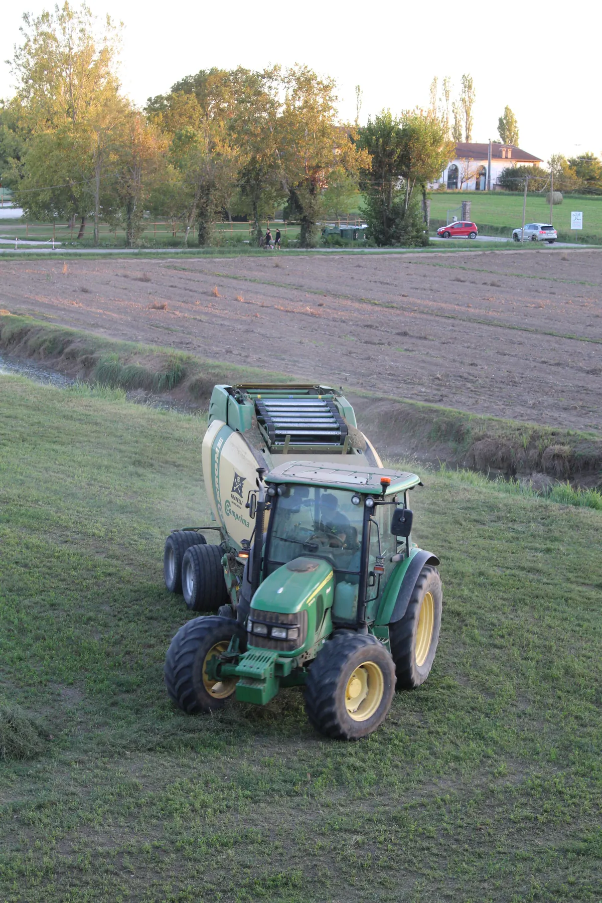 Trattore verde con la rotopressa visto dall’alto, mentre entra nel campo; dietro la terra arata, gli alberi e una casa