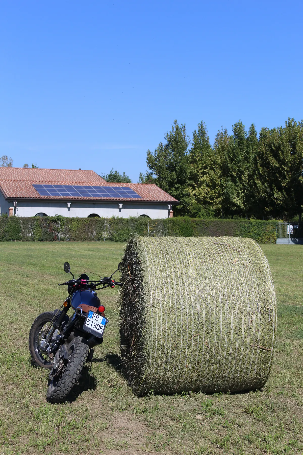 Il Tracker, una moto blu, appoggiato a una grande rotoballa di fieno in pieno sole, con la casa e i pannelli fotovoltaici dietro